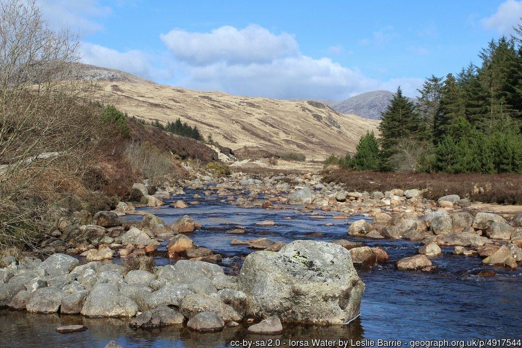 A natural scene with a river in the centre, heather and coniferous trees on the right and hills to the back.