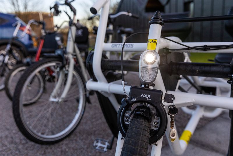 A row of bikes. A white bike is close to the camera, the light at the front is on as the day gets darker and comes towards evening.