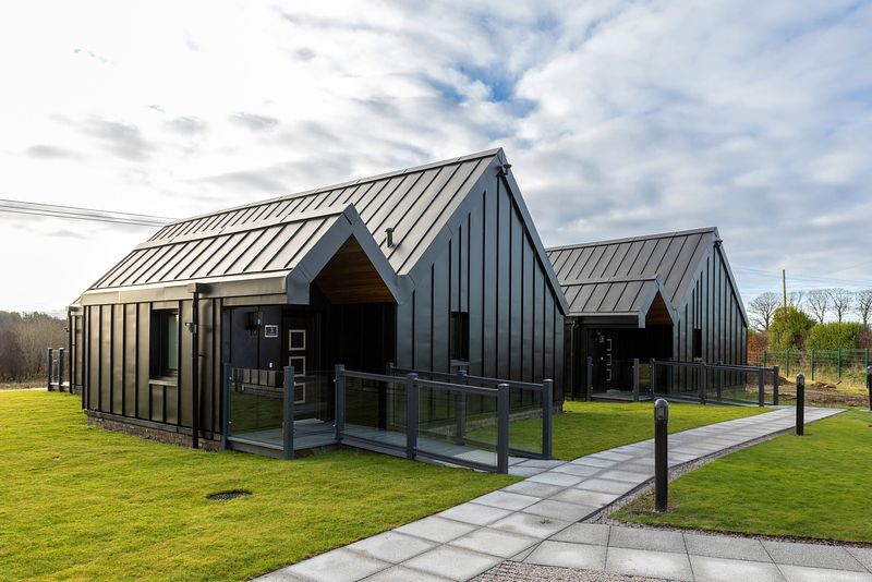 The chalets are standing in the sunlight on an area of short neat grass. This is the back view. There is a path made of grey paving stones connecting the entrances of the chalets and leading away out of shot.