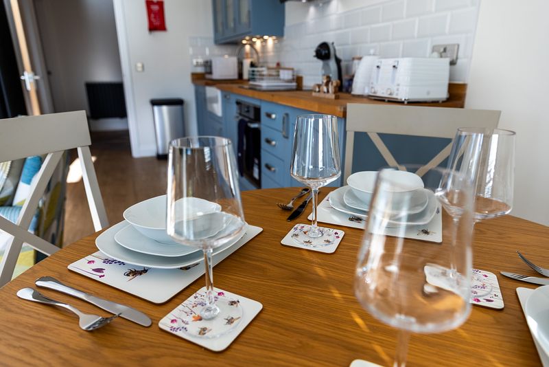 In the foreground a table is set with shiny clean glasses, nature themed placemats and tasteful plates and cutlery. In the background the kitchen can be seen which is part of the same room.