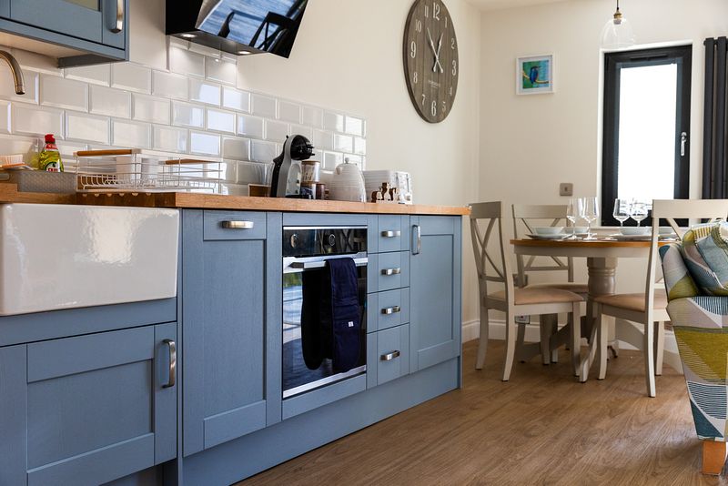 Modern and attractive kitchen and dining area with cabinets in a calm blue. There is a large clock on the wall and natural light streaming in a window to the back of the scene.