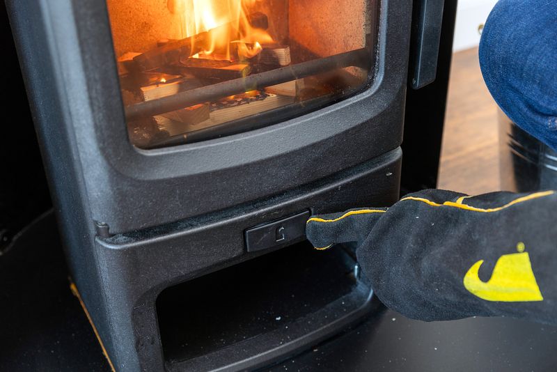 The stove is closed, and the pieces of wood are beginning to burn. A gloved hand is pointing at and just touching the air valve at the bottom of the stove.