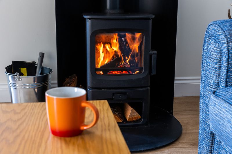 A wood burning stove is lit, and logs can be seen burning within. There is a blue seat to the right, a grey bucket with gloves and a tool handle protruding on the left. A cup sits on a table in the foreground.