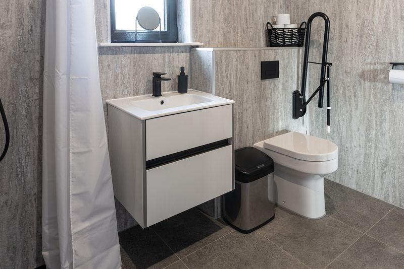 A modern and clean bathroom, featuring toilet and sink. The room is mostly a stylish grey with black highlights such as taps and soap dispenser. There is a handrail next to the toilet.